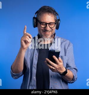 Glücklicher Mann mittleren Alters mit grauem Bart und Brille, der Musik mit kabellosen Kopfhörern genießt, während er ein Smartphone auf blauem Hintergrund verwendet Stockfoto