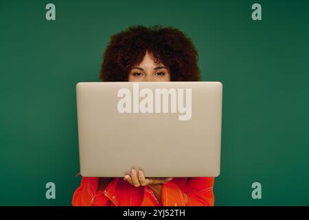 Eine junge afroamerikanische Frau strahlt vor Freude und zeigt ihr lockiges Haar, während sie einen Laptop in legerer Kleidung hält. Stockfoto