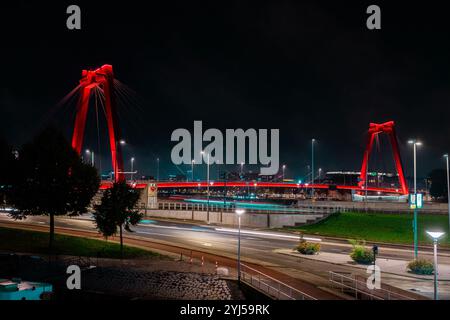 Willemsbrug Rotterdam bei Nacht, rot beleuchtet mit markanten Doppelmasten. Lichtspuren aus dem Verkehr schaffen eine dramatische urbane Szene. Stockfoto