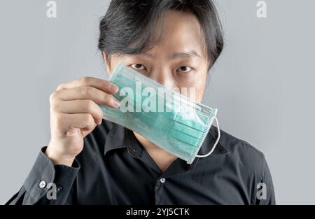 Asiatischer Mittelmann in einem schwarzen Hemd, trägt eine grüne Hygienemaske und kreuzt die Rückseite des Armes, steht vor einem weißen klaren Hintergrund. Stockfoto