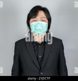 Asiatischer Mittelmann in einem schwarzen Hemd, trägt eine grüne Hygienemaske und kreuzt die Rückseite des Armes, steht vor einem weißen klaren Hintergrund. Stockfoto