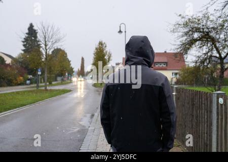 Langweid, Bayern, Deutschland - 13. November 2024: Symbolisches Bild von Wetterwechsel und Schneefall in Bayern. Ein Mann mit Kapuze und Winterjacke geht bei bewölktem Wetter *** Symbolbild Wetterumschwung und Schneefall in Bayern. Ein Mann mit Kaputze und Winterjacke läuft bei trübem Wetter spazieren Stockfoto