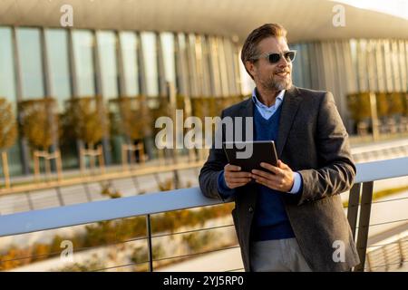 Ein gut gekleideter Mann lächelt, während er ein Tablet auf einem Balkon benutzt, umgeben von zeitgenössischer Architektur und lebhaftem Herbstlaub, und genießt einen Moment Stockfoto