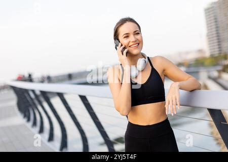 Eine Joggerin hält an ihrem Telefon am Fluss an, trägt Trainingsausrüstung und Kopfhörer und genießt die warme Sonne und die ruhige Atmosphäre Stockfoto