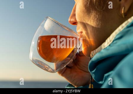 Frau genießt heißes Getränk in doppelwandigem Glas mit Meerblick bei Sonnenuntergang Stockfoto