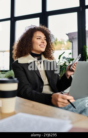 Eine Frau mit lockigen Haaren lächelt, während sie auf ihr Handy schaut, während sie an ihrem Laptop arbeitet. Stockfoto