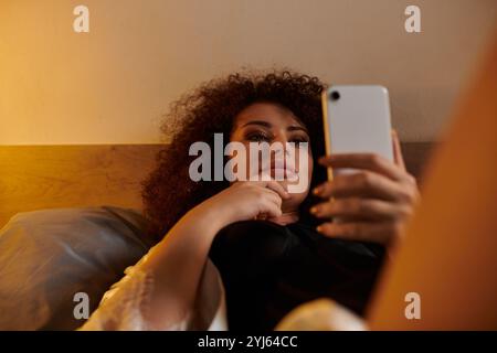 Eine Frau, die sich im Bett entspannt, durchblättert ihr Smartphone, in Gedanken verloren. Stockfoto