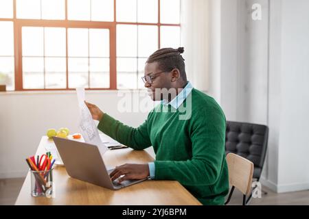 Ein schwarzer Mann mit Brille sitzt an einem hölzernen Schreibtisch in einem hellen Bürobereich. Er konzentriert sich auf die Überprüfung von Dokumenten, während er an einem Laptop arbeitet, umgeben von Stockfoto