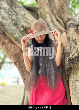 Eine Frau mit geradem, langem Haar, die einen Hut hält, der sich auf einen großen Baumstamm in einem tropischen Land stützt. Stockfoto