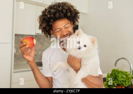 Ein schwarzer Mann lächelt breit in einer hellen Küche, hält einen Apfel in der einen Hand und einen flauschigen weißen Hund in der anderen. Frische Produkte sind auf dem Coun sichtbar Stockfoto