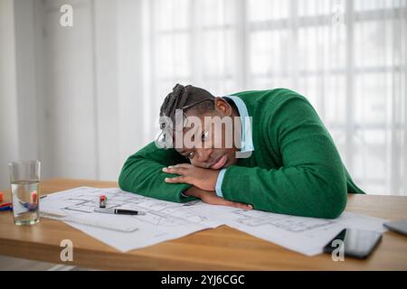 Ein junger schwarzer Mann mit Brille scheint müde und frustriert zu sein, während er seinen Kopf auf einem überfüllten Schreibtisch mit Blaupausen und Schreibwaren in einem Brunnen ruht Stockfoto