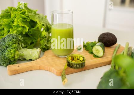 Ein leuchtend grüner Smoothie sitzt in einem Glas auf einem Schneidebrett, umgeben von frischem Brokkoli, Avocado, Salat, Gurke und Maßband. Hervorhebung Stockfoto