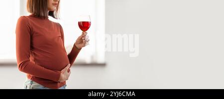 Eine junge Frau steht in einem hellen Innenraum und legt sich sanft in die Wiege ihres Babys. Sie hält ein Glas, gefüllt mit einer roten Flüssigkeit, reflektiert einen Moment der Ruhe Stockfoto