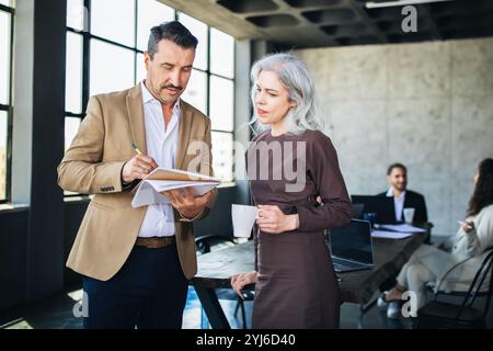 Zwei Kollegen führen eine konzentrierte Diskussion, während sie sich in einem modernen Büro Notizen ansehen. Andere arbeiten im Hintergrund und tragen zu einem Kollabo bei Stockfoto