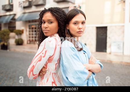 Zwei junge Frauen posieren selbstbewusst auf einer charmanten Straße. Einer hat lockige Haare und ein buntes Outfit, der andere trägt ein hellblaues Oberteil, b Stockfoto