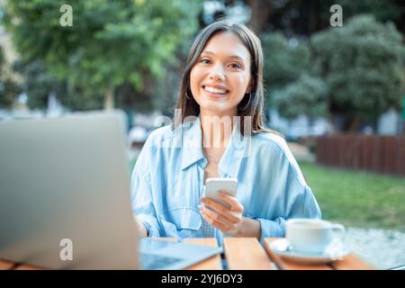 Eine junge Frau lächelt, während sie an einem Café-Tisch im Park sitzt, ihr Smartphone hält und ihren Laptop benutzt. Die Umgebung im Freien bietet eine ruhige Atmosphäre Stockfoto