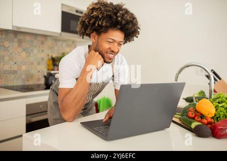 Ein schwarzer Mann mit lockigen Haaren lächelt, als er ein Rezept auf seinem Laptop in einer modernen Küche folgt. Frisches Gemüse ist in der Nähe ordentlich angeordnet und zeigt ein Stockfoto