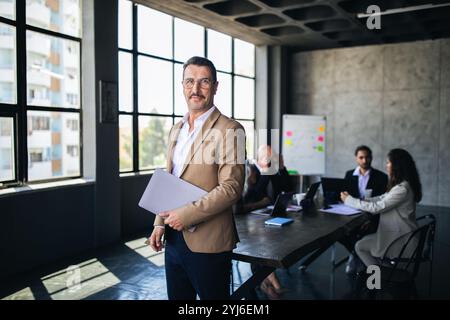 Ein Mann in einem beigefarbenen Blazer steht selbstbewusst in einem modernen Büro und hält Dokumente. Hinter ihm diskutieren drei Kollegen an einem großen Tisch i Stockfoto