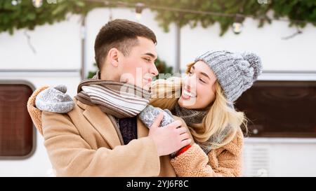 Ein lächelndes Paar umarmt sich herzlich, während es eine Winterfeier genießt, gekleidet in gemütliche Tücher und Hüte, umgeben von festlichen Dekorationen und einem Cheerf Stockfoto