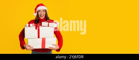 Eine fröhliche Frau in rotem Pullover und Weihnachtsmann-Hut trägt freudig eingewickelte Weihnachtsgeschenke. Der hellgelbe Hintergrund trägt zur festlichen Atmosphäre bei Stockfoto