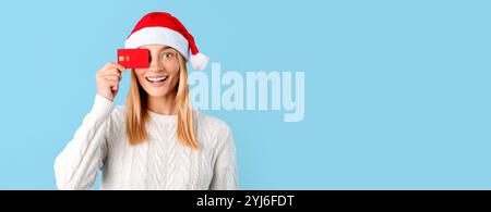 Aufgeregte europäische Dame im Weihnachtsmann-Hut bedeckt Auge mit Kreditkarte isoliert auf blauem Hintergrund, Studio-Porträt. Frohe Neujahrsfeier Stockfoto