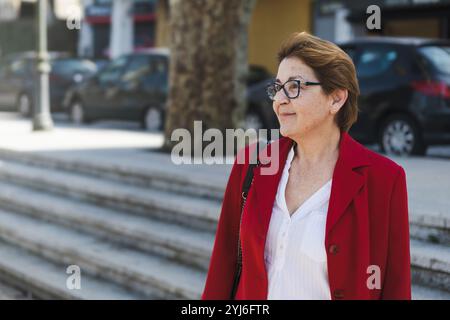 Eine reife Frau mit roter Jacke und Brille lächelt sanft, während sie auf einem Platz einer Stadt steht und einen ehrlichen Moment des Stadtlebens festhält. Stockfoto