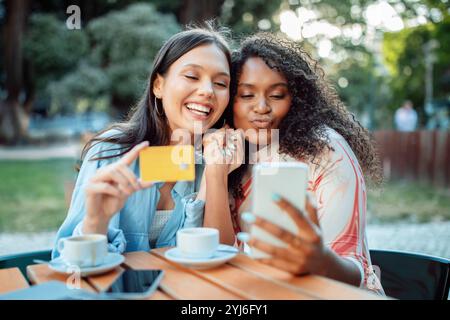Zwei Freunde sitzen an einem Holztisch, lächeln und machen zusammen ein Selfie. Sie haben eine Kreditkarte und ein Telefon, während sie Kaffee in einem lebhaften Park A genießen Stockfoto