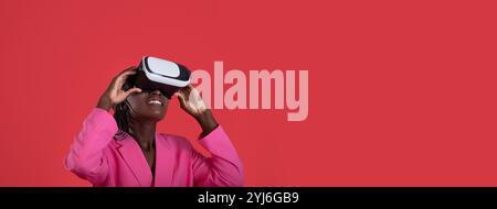 Eine junge schwarze Frau in einem rosa Blazer lächelt, während sie sich mit einem Headset in eine virtuelle Realität eintaucht. Der leuchtende rote Hintergrund Stockfoto