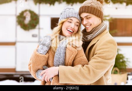 Ein fröhliches Paar teilt sich in Winterkleidung in einer verschneiten Landschaft eine warme Umarmung. Sie lächeln hell und genießen die gemütliche Atmosphäre der kalten Jahreszeit Stockfoto