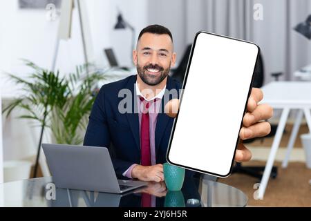 Ein Mann in formeller Kleidung sitzt an einem Schreibtisch, lächelt und hält ein Smartphone vor sich. Sein Laptop und eine Tasse sind in der Nähe, was ein produktives Produkt schafft Stockfoto