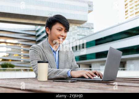 Ein junger asiatischer Mann, der in einem schicken Blazer gekleidet ist, konzentriert sich auf seinen Laptop, während er an einem Holztisch vor einem modernen Gebäude unter dem After sitzt Stockfoto