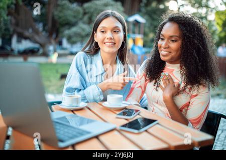 Zwei Freunde sitzen an einem Holztisch in einem lebhaften Café, lächeln und unterhalten sich, während sie einen Kaffee genießen. Laptops und Telefone sind um sie herum und schaffen eine entspannte Atmosphäre Stockfoto