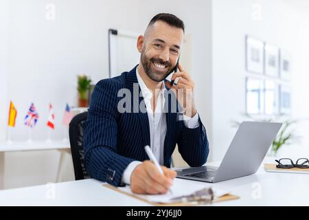 Ein gut gekleideter Mann sitzt an einem Schreibtisch und lächelt, während er telefoniert. Er schreibt Notizen auf eine Zwischenablage, vor ihm ist ein Laptop offen und die Flaggen von va Stockfoto