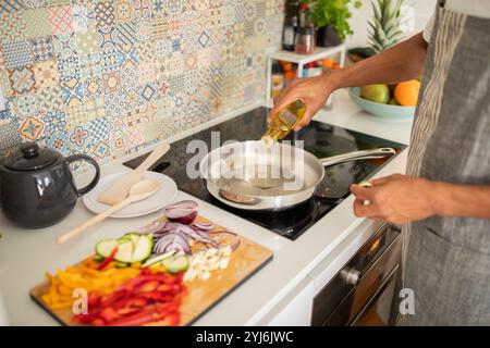 Eine Person bereitet sich zum Kochen vor, indem sie Öl in eine Pfanne gießt. Frisches Gemüse wie Paprika, Zwiebeln und Zucchini wird auf einem Schneidebrett geschnitten Stockfoto