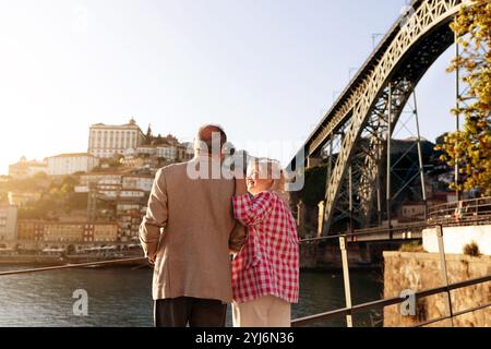 Ein Paar steht zusammen an einer Uferpromenade und bewundert den Sonnenuntergang, während das warme Licht die historischen Gebäude der Stadt und die imposante Brücke beleuchtet Stockfoto