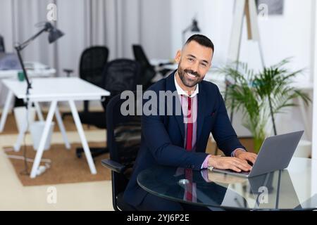 Ein gut gekleideter Mann sitzt an einem Glasschreibtisch in einer modernen Büroumgebung und beschäftigt sich mit seinem Laptop. Der Arbeitsbereich verfügt über ergonomische Stühle und greene Stockfoto