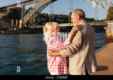 Ein reifes Paar teilt sich einen intimen Moment am Fluss, umhüllt und lächelt sich gegenseitig an. Die Umgebung bietet eine wunderschöne Brücke im Hintergrund d Stockfoto