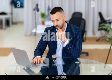Ein fokussierter Mann im Anzug sitzt an einem Glasschreibtisch und benutzt in einem modernen Büro einen Laptop. Er hält eine kleine Tasse Kaffee, während er tief nachdenkt. Stockfoto
