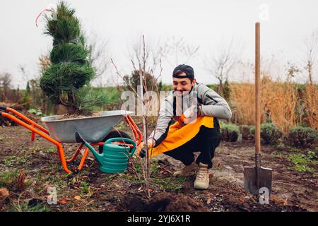 Gärtner pflanzt Bäume im herbstlichen Garten mit Schubkarre, Schaufel und Gießkanne. Saisonale Arbeiten im Herbstgarten. Stockfoto