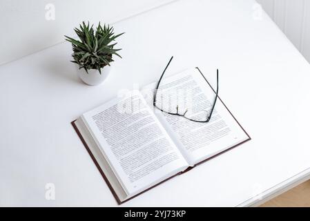 Brille auf offenem Buch mit dichtem Text, symbolisiert Lernen, Lesen und Wissenserwerb; minimalistischer Schreibtischbereich mit einer Pflanze fügt hinzu Stockfoto