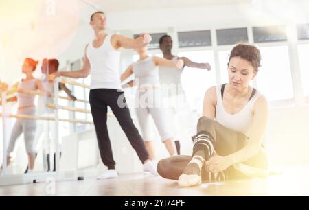 Die Ballerina bindet ihre spitzenschuhe während der Pause zwischen den Ballettproben an die Füße. Andere Teilnehmer führen weiterhin verschiedene Ballettschritte durch Stockfoto