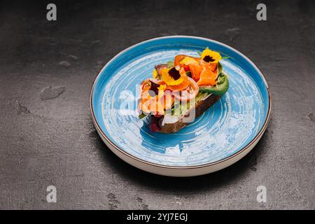 Gourmet-Toast mit geräuchertem Lachs mit frischem Gemüse und essbaren Blumen auf einem leuchtend blauen Teller Stockfoto