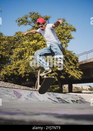 Skateboarder in Red Cap Sprüngen im sonnigen Skatepark umgeben von Bäumen. Stockfoto
