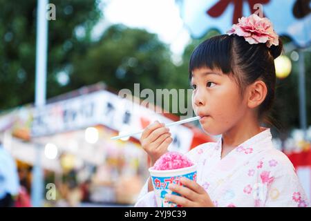 Ein Mädchen in Yukata isst rasiertes Eis, das von einem Festivalstand gekauft wurde Stockfoto