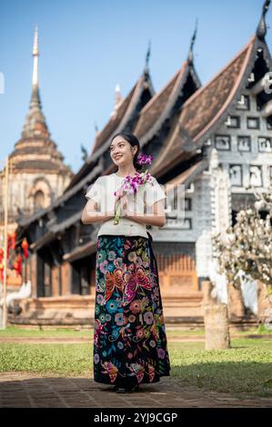 Eine charmante, lächelnde asiatische Frau in einem traditionellen thailändischen Lanna-Kleid steht draußen mit einem wunderschönen alten Tempel und einer Pagode im Hintergrund, Holdin Stockfoto