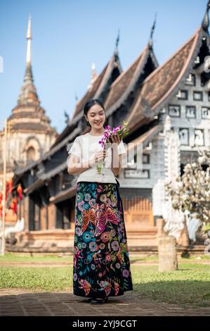 Eine charmante, lächelnde asiatische Frau in einem traditionellen thailändischen Lanna-Kleid steht draußen mit einem wunderschönen antiken Tempel und einer Pagode im Hintergrund, Admiri Stockfoto