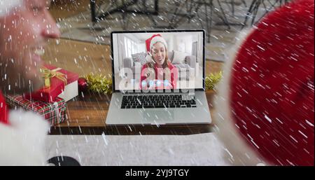 Bild von Schnee, der über ein lächelndes Paar in santa-Hüten fällt, während eines Laptop-Videogesprächs mit der Familie. weihnachten, Tradition und Festkonzept digital generativ Stockfoto