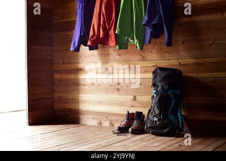 Vier Paar Bergsteigerbekleidung in vier verschiedenen Farben, die an Haken in der Hütte hängen, Schuhe und Rucksack auf dem Boden Stockfoto