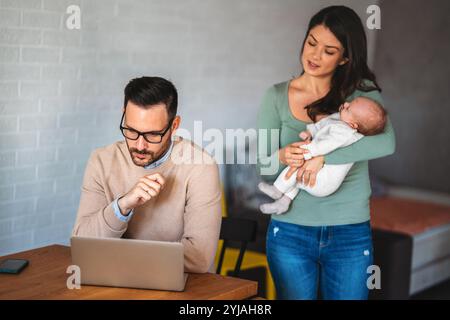 Der junge Vater arbeitet am Laptop, während seine Frau sich um ein Kleinkind kümmert. Arbeiten Sie von zu Hause aus Stockfoto