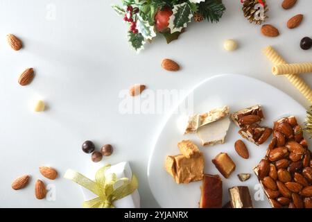 Teller mit verschiedenen Portionen Nougat auf weißem Tisch mit natürlichen Mandeln, Schokoladenkugeln und Weihnachtsdekoration. Draufsicht. Stockfoto